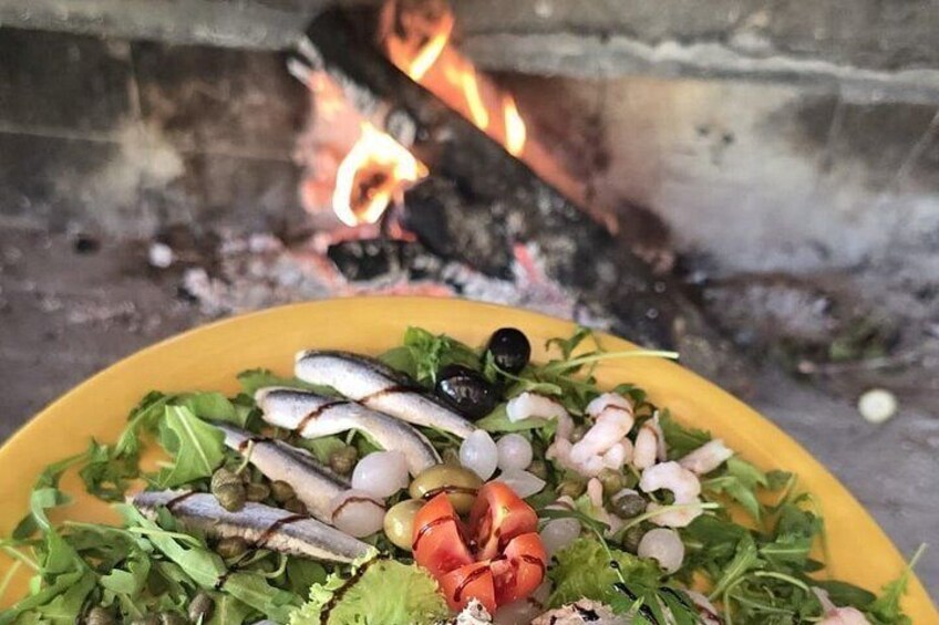 A dish of seafood with greens and a red pepper, set against a backdrop of a wood-fired oven. 5