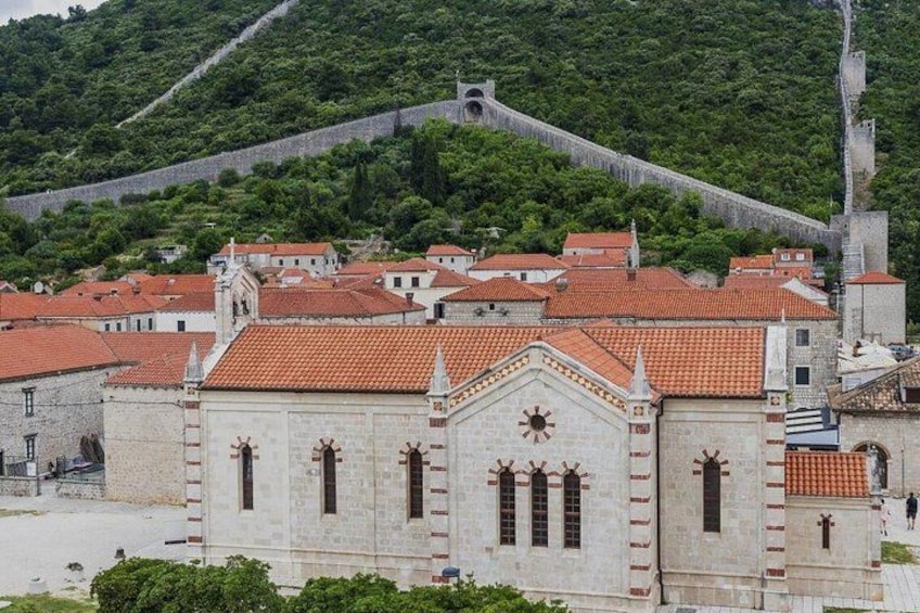 A historic town with red-tiled roofs, a prominent church with a cross on its facade, and a fortified wall in the background. 3