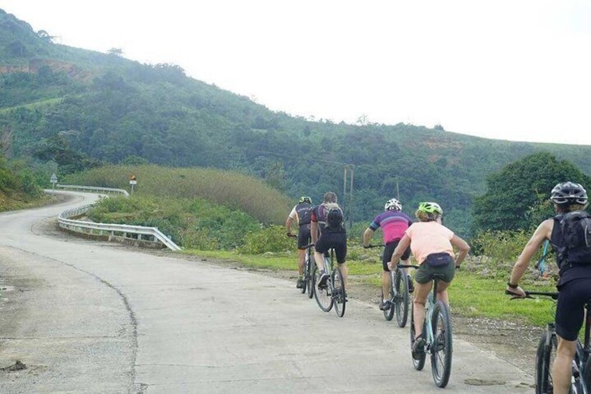 A group of cyclists on a paved road with a guardrail, surrounded by lush greenery and hills. 4