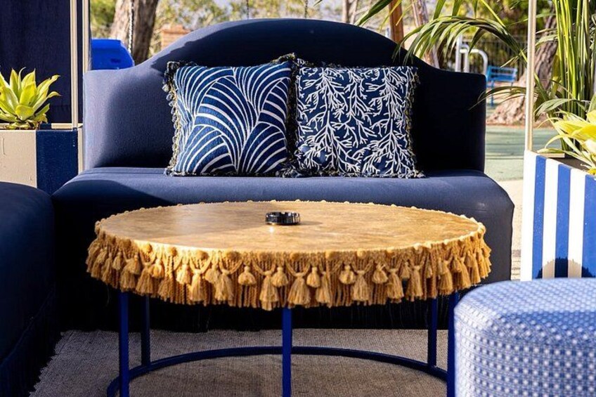 A round wooden table with a fringe edge, a dark blue sofa with patterned cushions, and a blue and white striped planter. 5