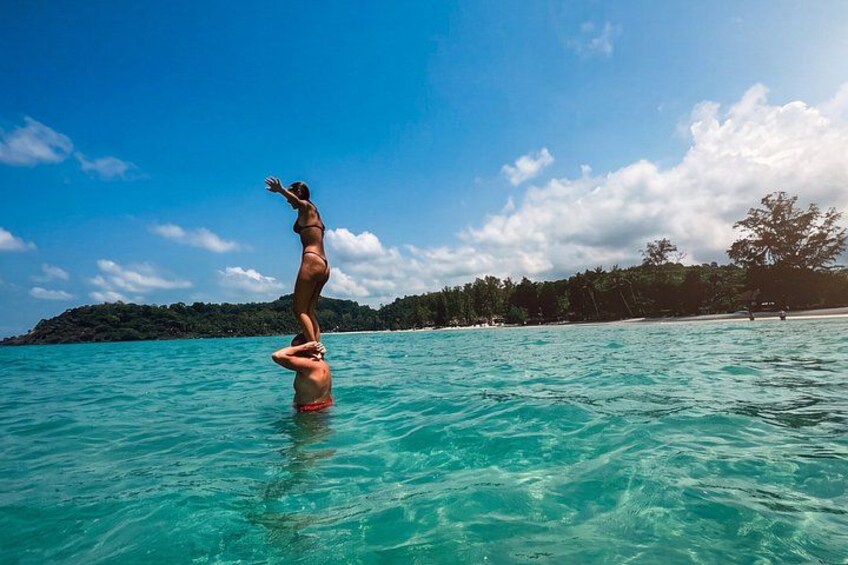 A person balancing on another person's shoulders in clear turquoise water with a beach and trees in the background. 3