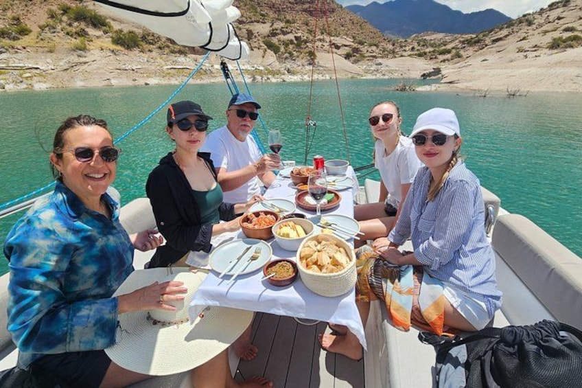 A group of people dining on a boat with a table set with food and drinks. 5