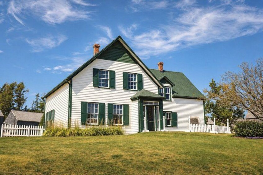 A two-story white house with green shutters and a green roof, surrounded by a white picket fence and a grassy lawn. 2