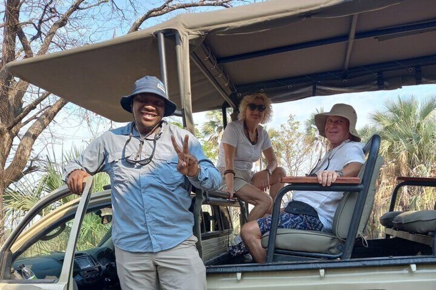A safari vehicle with three people, one standing and two seated, in a natural setting with trees and palm trees. 5