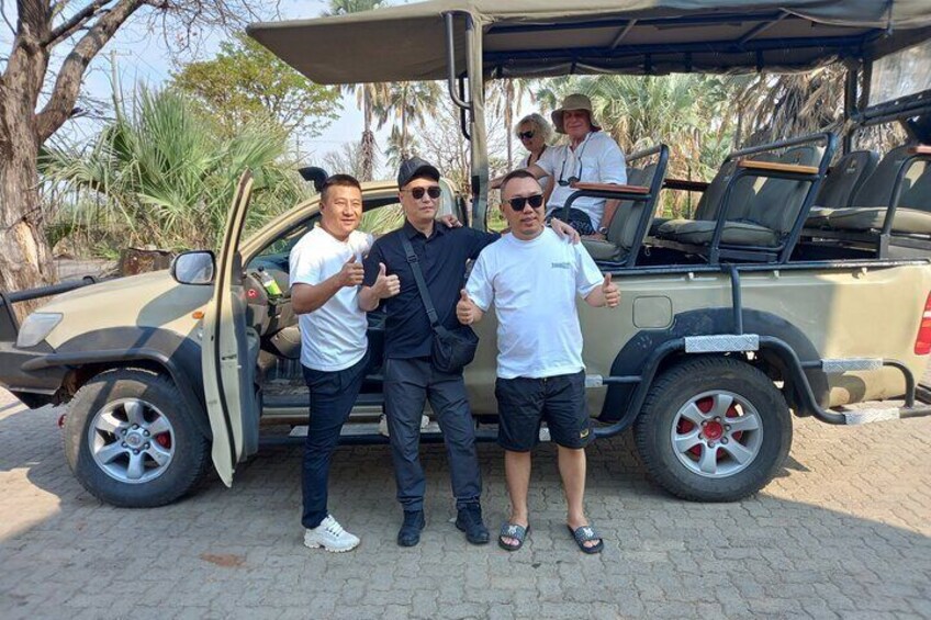 A group of people standing next to an open safari vehicle with a roof, in a natural setting with trees and a clear sky. 4
