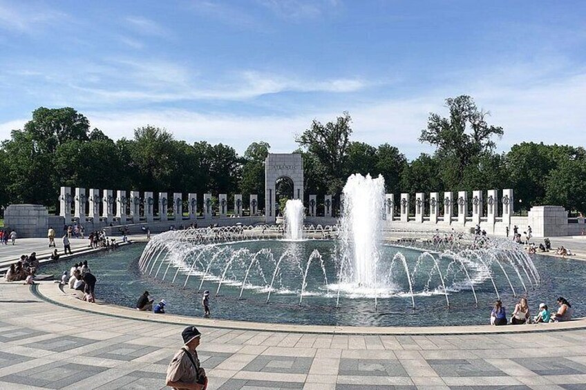 A large fountain with multiple water jets, surrounded by a paved area with people sitting and walking. In the background, there is a monument with columns and a clear sky. 4