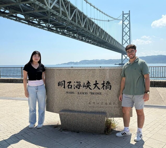 Two people standing in front of a stone monument with Japanese text, under a bridge with a view of the sea and mountains in the background. 2