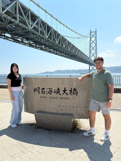 A man and a woman standing in front of a stone marker with text, under a bridge with a clear view of the sea and distant mountains. 4
