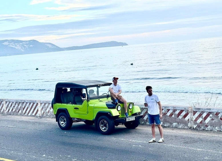 A green vehicle parked by the roadside with a body of water and mountains in the background. 5