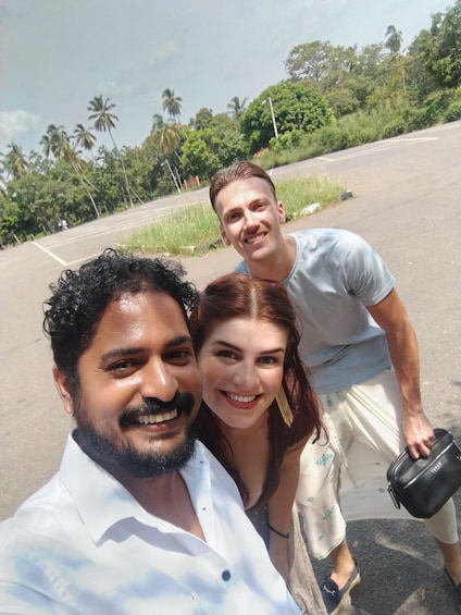 Trois personnes prennent un selfie sur une route, avec une végétation luxuriante et un ciel dégagé en arrière-plan. 3