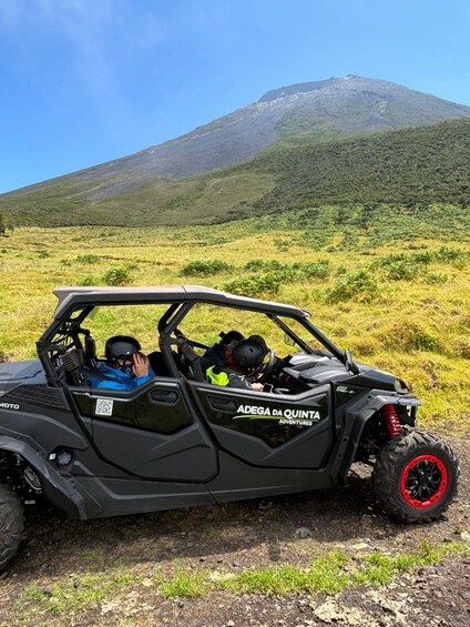 A black off-road vehicle with 'ADEGA DA QUINTA ADVENTURES' on the side parked on a dirt road with a grassy field and a mountain in the background. 3