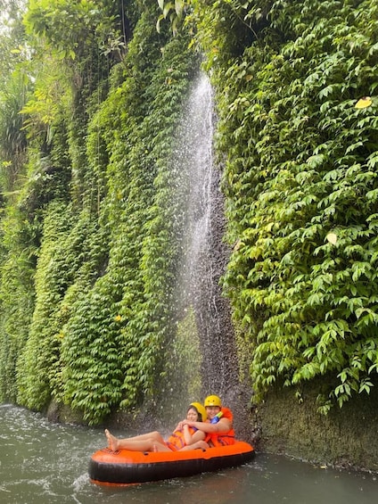 Dos personas en una balsa naranja flotando cerca de una cascada rodeada de frondosa vegetación verde. 4