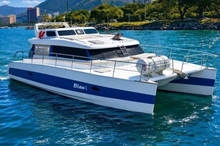 A white and blue catamaran named 'Blam' on calm waters with a cityscape in the background. 1