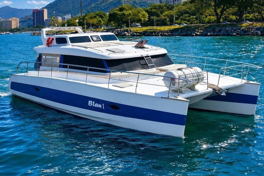 A white and blue catamaran named 'Blam!' on calm water with a cityscape and greenery in the background. 2