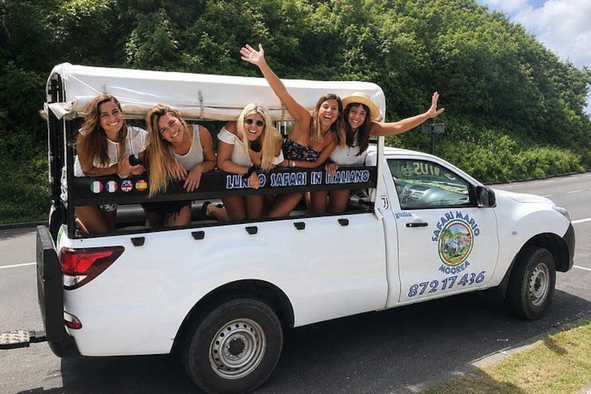 A white pickup truck with a group of people on the back, parked on a road with greenery in the background. 4