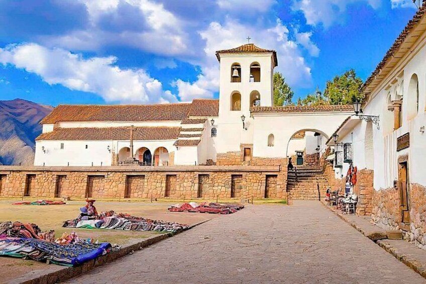 A plaza with a white church, a bell tower, and a market with colorful textiles. 1