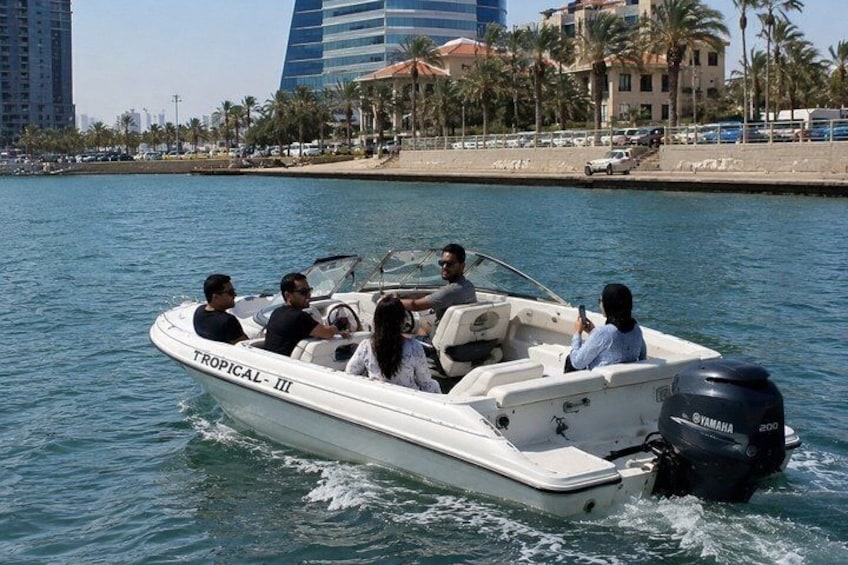 A white motorboat named 'Tropical III' on water with people on board, a cityscape with tall buildings in the background, and a sandy beach with palm trees. 2