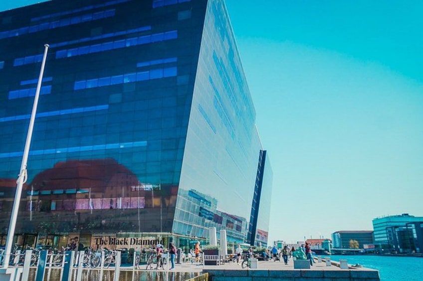 A modern glass building with reflections, a clear blue sky, and a busy waterfront with people and bicycles. 3