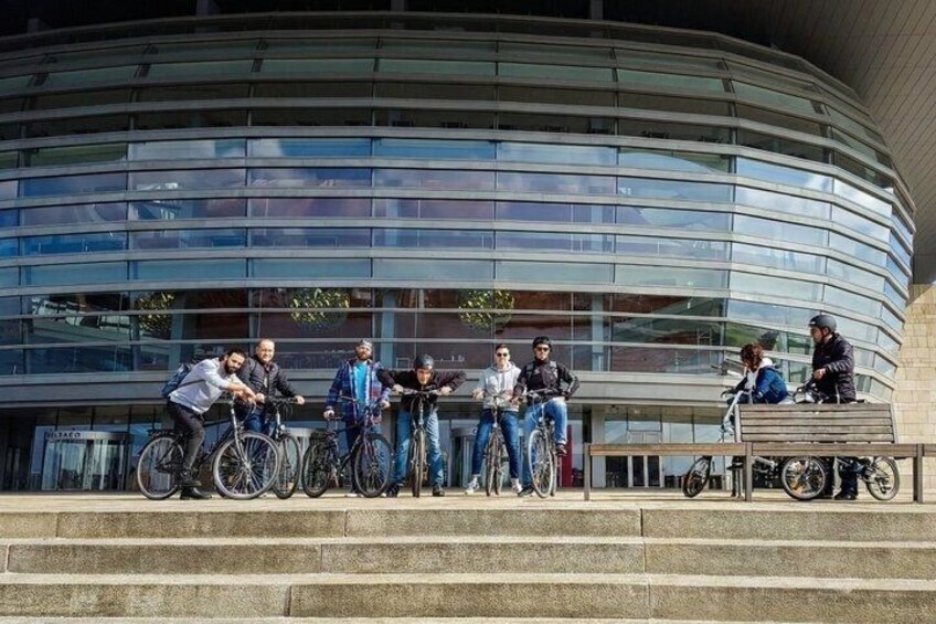 A group of people with bicycles in front of a modern building with a curved glass facade. 4