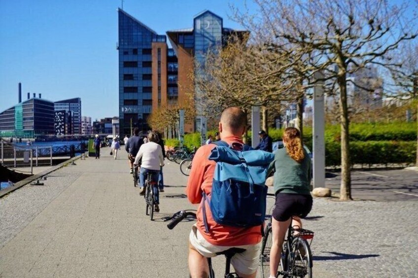 A paved walkway with cyclists, a river, modern buildings, and trees. 5