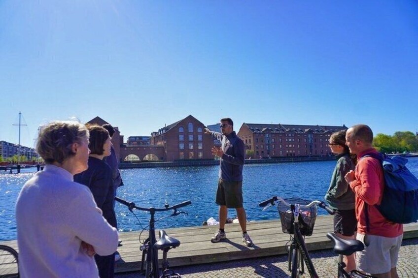 A group of people standing by a waterfront with bicycles, a dock, and buildings in the background. 1