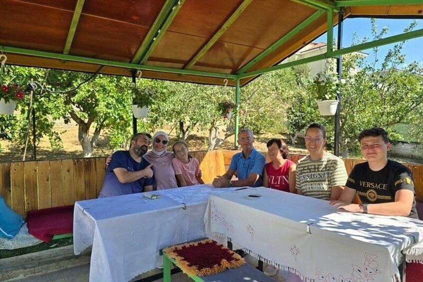 A group of people seated at a table under a covered area with a tablecloth, surrounded by trees. 3