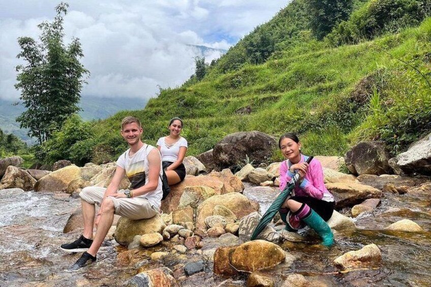 Three people sitting on rocks by a stream in a mountainous area. 3