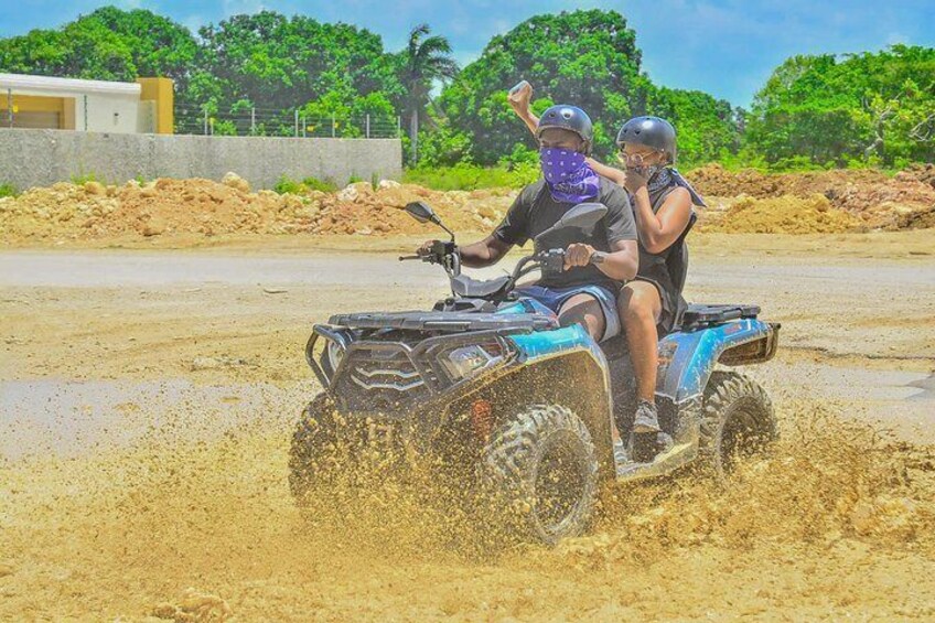 Two people on an ATV in a muddy area with trees and a building in the background. 4