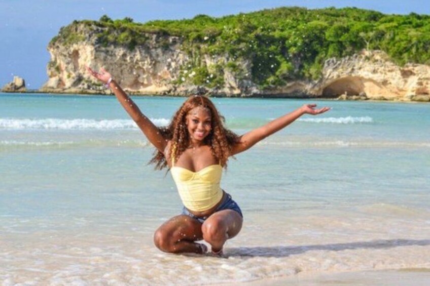A person squatting on a beach with arms raised, facing a cliff with greenery and the sea. 2