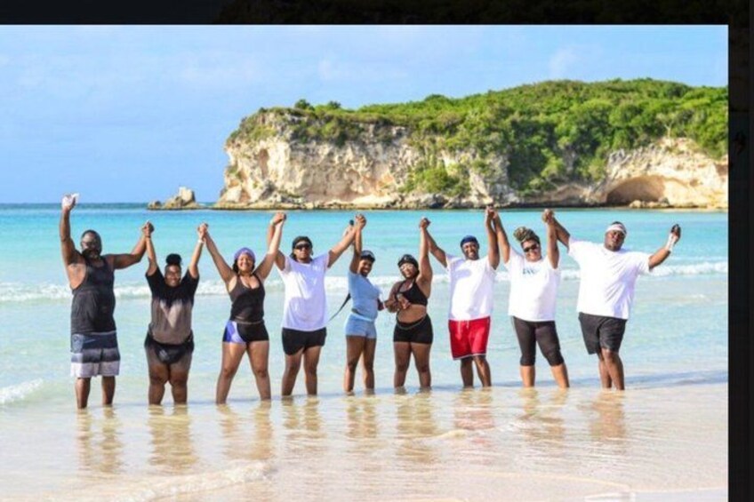 A group of people standing on a beach with their arms raised, facing a cliff with greenery. 5