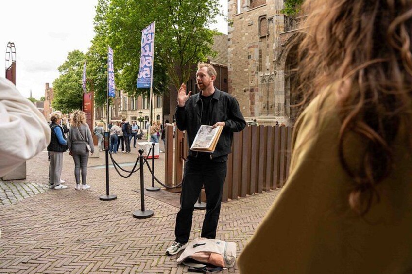 A person presenting with a framed item, standing on a brick-paved street with buildings and flags in the background. 5
