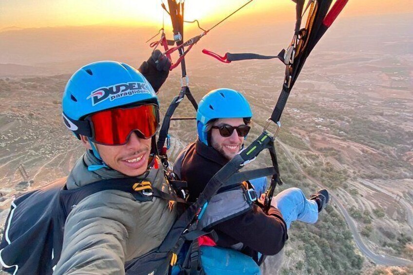 Two individuals in a tandem paragliding harness with a clear sky and a landscape below. 2