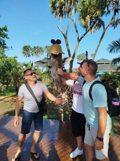 Three people interacting with a giraffe wearing a hat, in a park with palm trees. 5