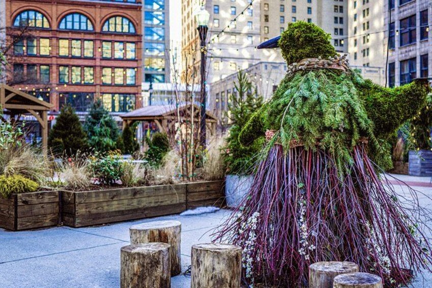 A moss-covered sculpture resembling a bird in an urban plaza with wooden stumps and a building in the background. 4