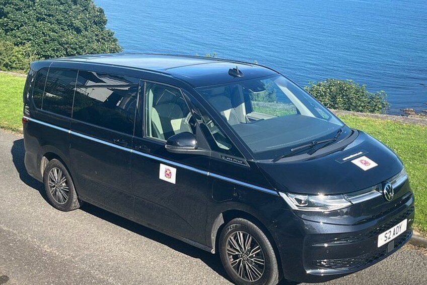 A black van parked on a road with a body of water and greenery in the background. 3