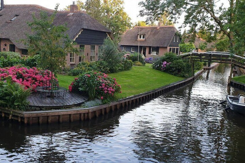 A canal with a wooden bridge, a house with a thatched roof, and a garden with pink flowers. 4