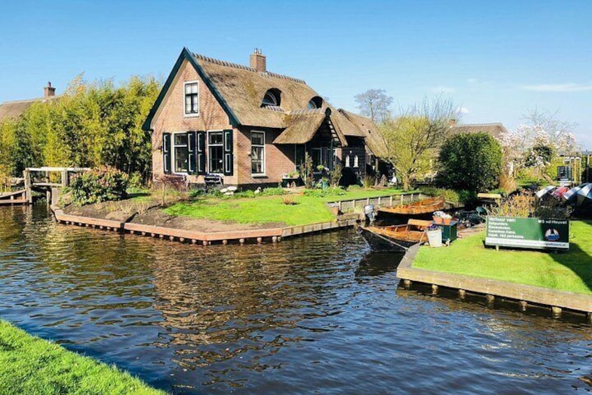 A traditional house by a canal with a thatched roof, surrounded by greenery and a small boat docked nearby. 3
