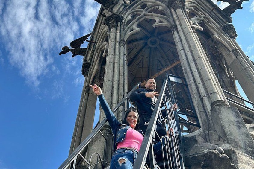 Two people climbing a gothic-style tower with intricate stonework and a clear blue sky in the background. 2