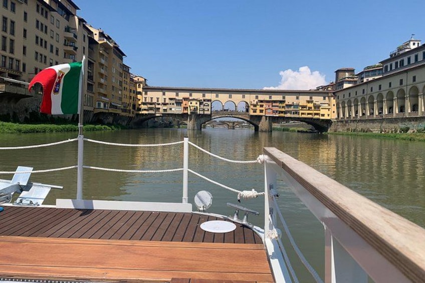 A boat deck with a flag, a bridge, and buildings in the background. 1