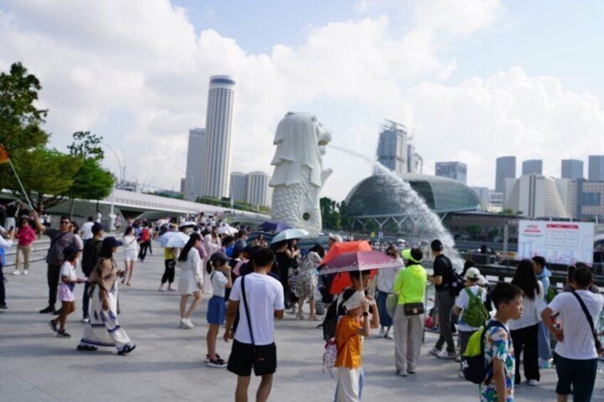 A crowded urban square with a large white statue and modern buildings in the background. 2