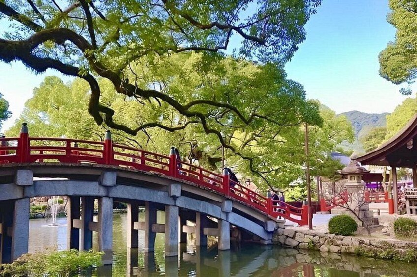 A stone bridge with red railings over a calm river, surrounded by lush greenery and a traditional building. 5