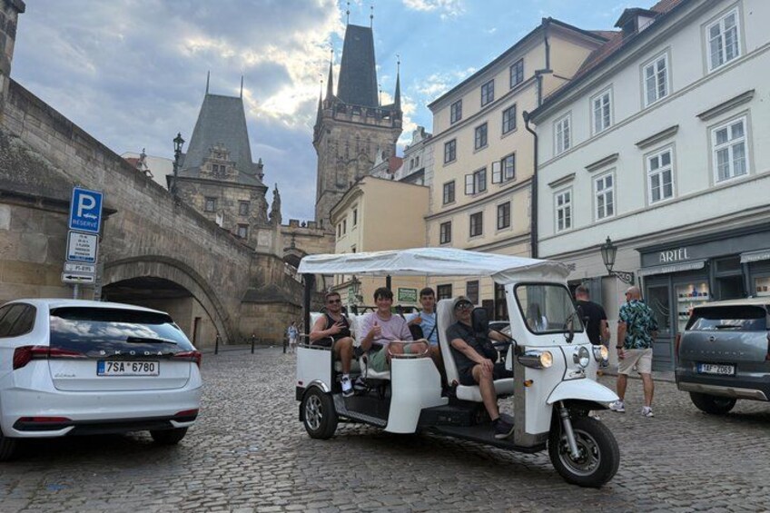 A white three-wheeled vehicle with passengers, parked on a cobblestone street, with historic buildings and a bridge in the background. 1