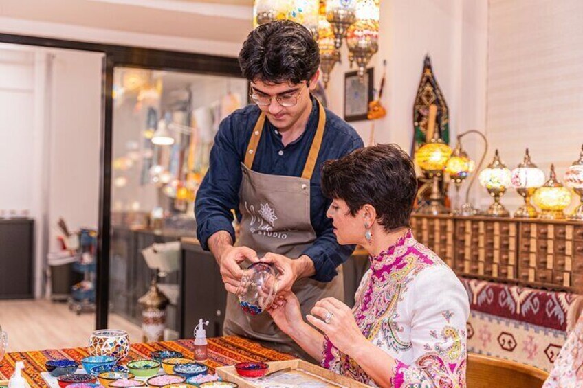 A person crafting colorful glass pieces at a table with various glass items and decorative lamps in the background. 3