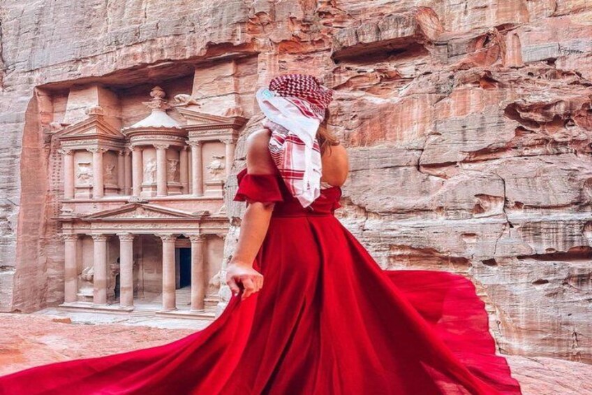 A person in a red dress standing in front of a carved rock facade with a building structure. 3