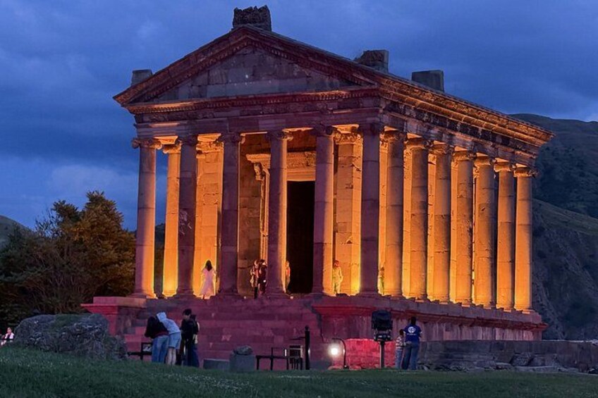 An ancient Greek temple with columns, illuminated by artificial lighting, surrounded by a grassy area with people. 1