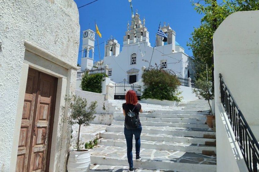 A person walking up a white stone staircase towards a white church with multiple bells and a blue dome. 1