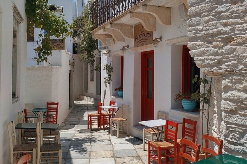 A narrow street with white buildings, red doors, and outdoor seating with red chairs and tables. 3