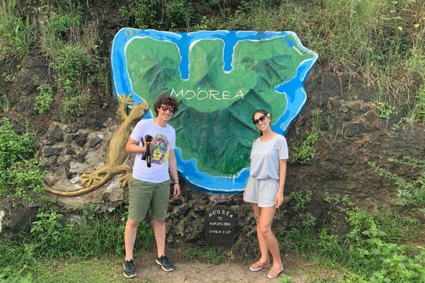 Two people standing in front of a large, colorful map with the word 'MOOREA' and a lizard graphic. 2