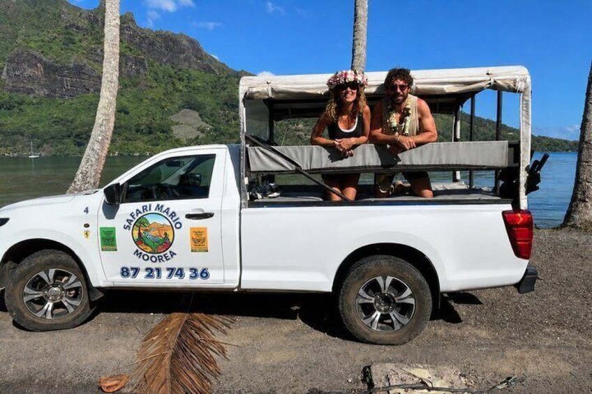 A white safari vehicle with a logo and contact number parked by a body of water with palm trees and a mountainous backdrop. 1