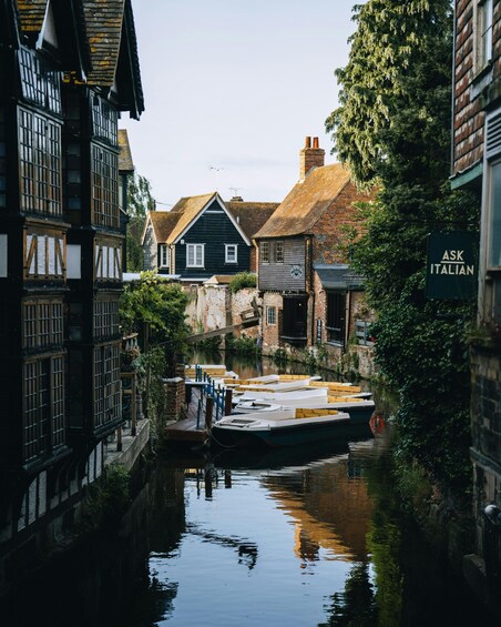 A narrow canal with boats docked, traditional half-timbered houses, and a sign for 'ASK ITALIAN.' 5